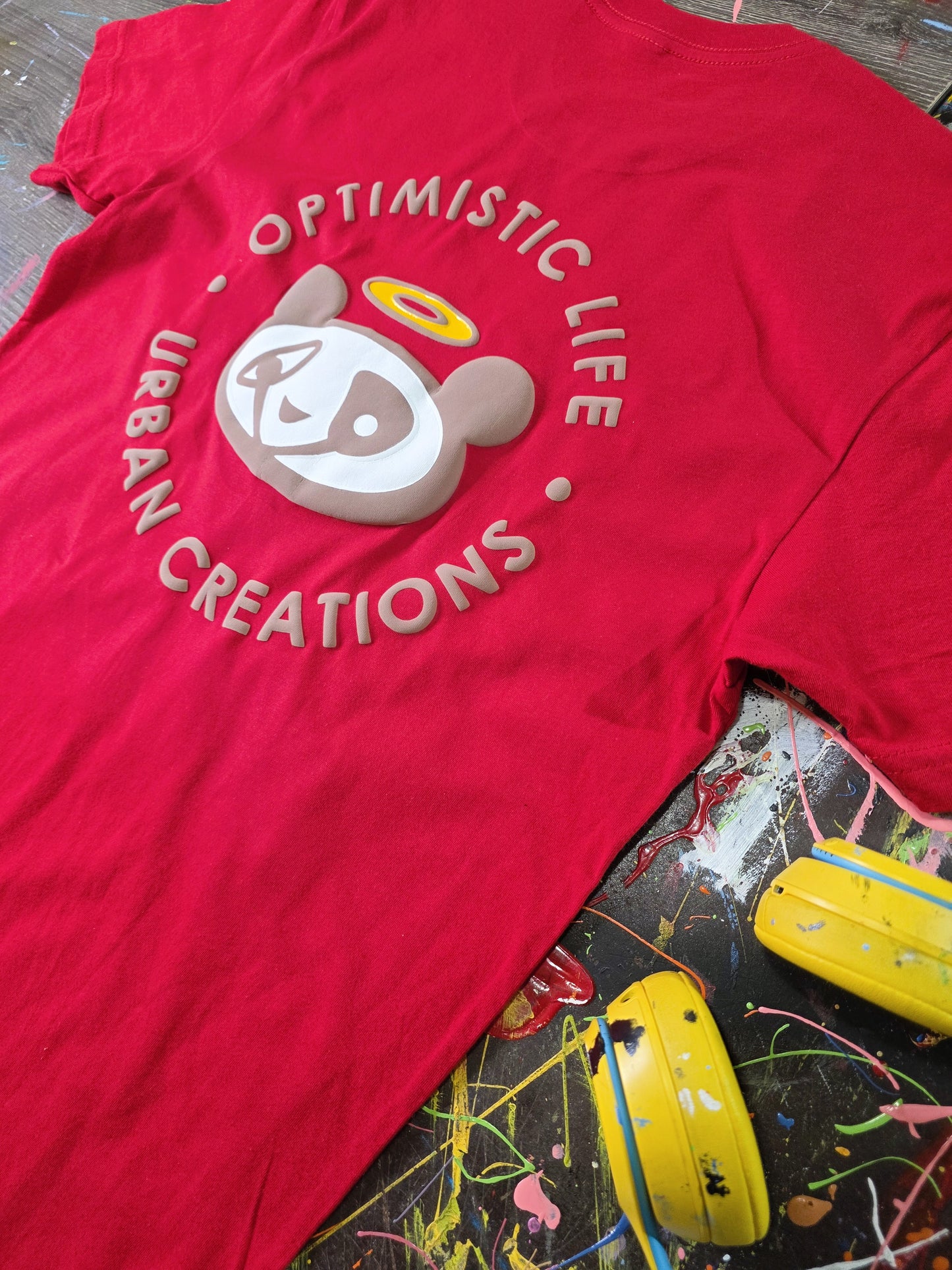 Red t-shirt with 'Optimistic Life Urban Creations' design on a colorful surface.